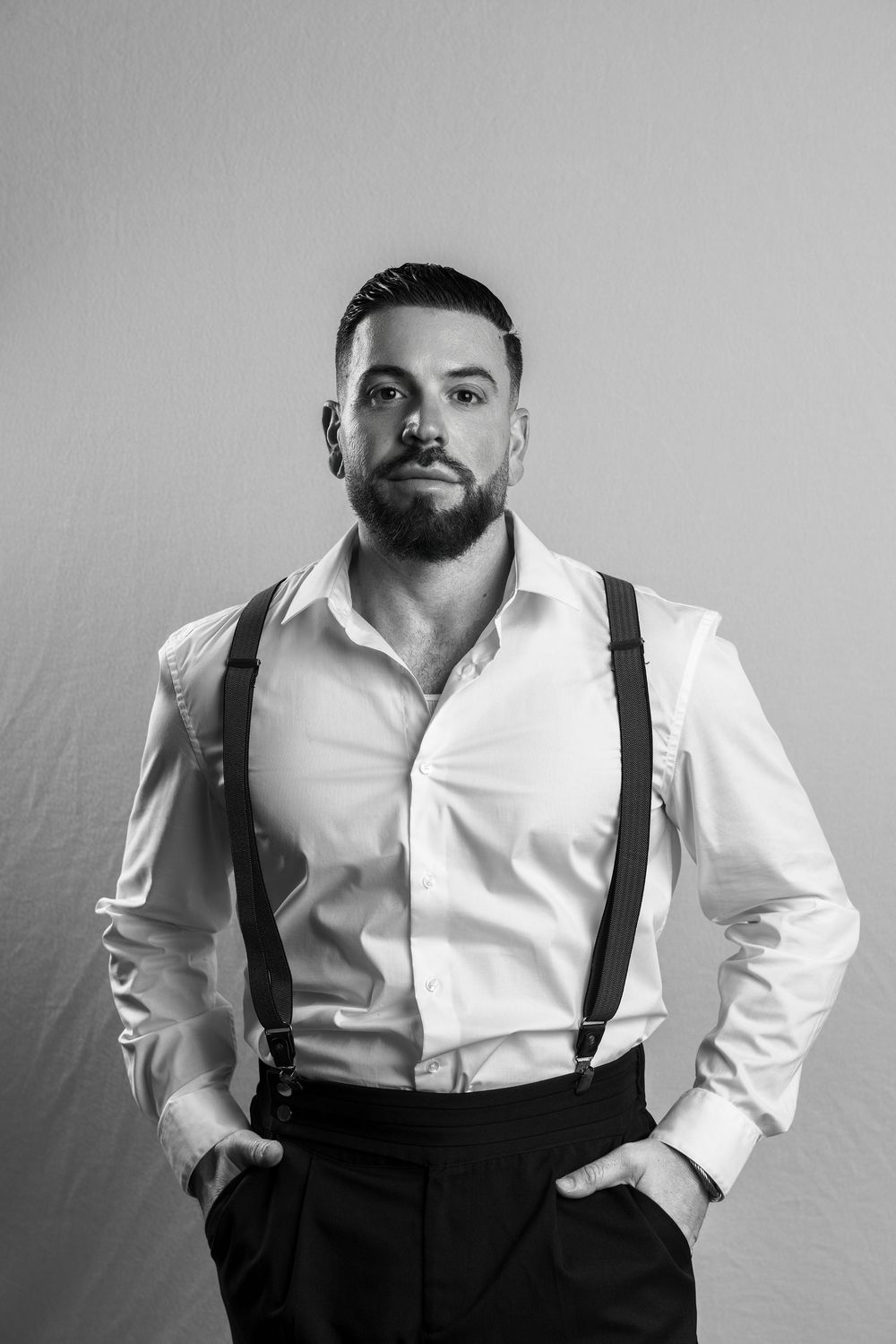 Black and white portrait of a bearded man in white dress shirt with suspenders, hands in pockets.