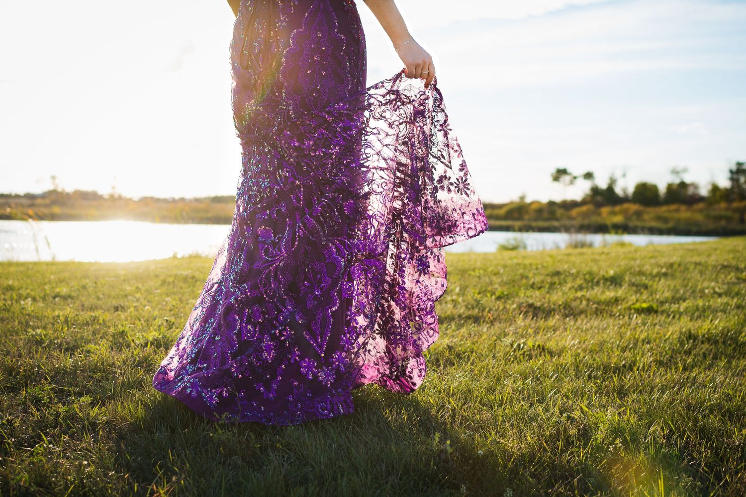 Purple lace dress flows in golden sunlight on grassy field near water at sunset.
