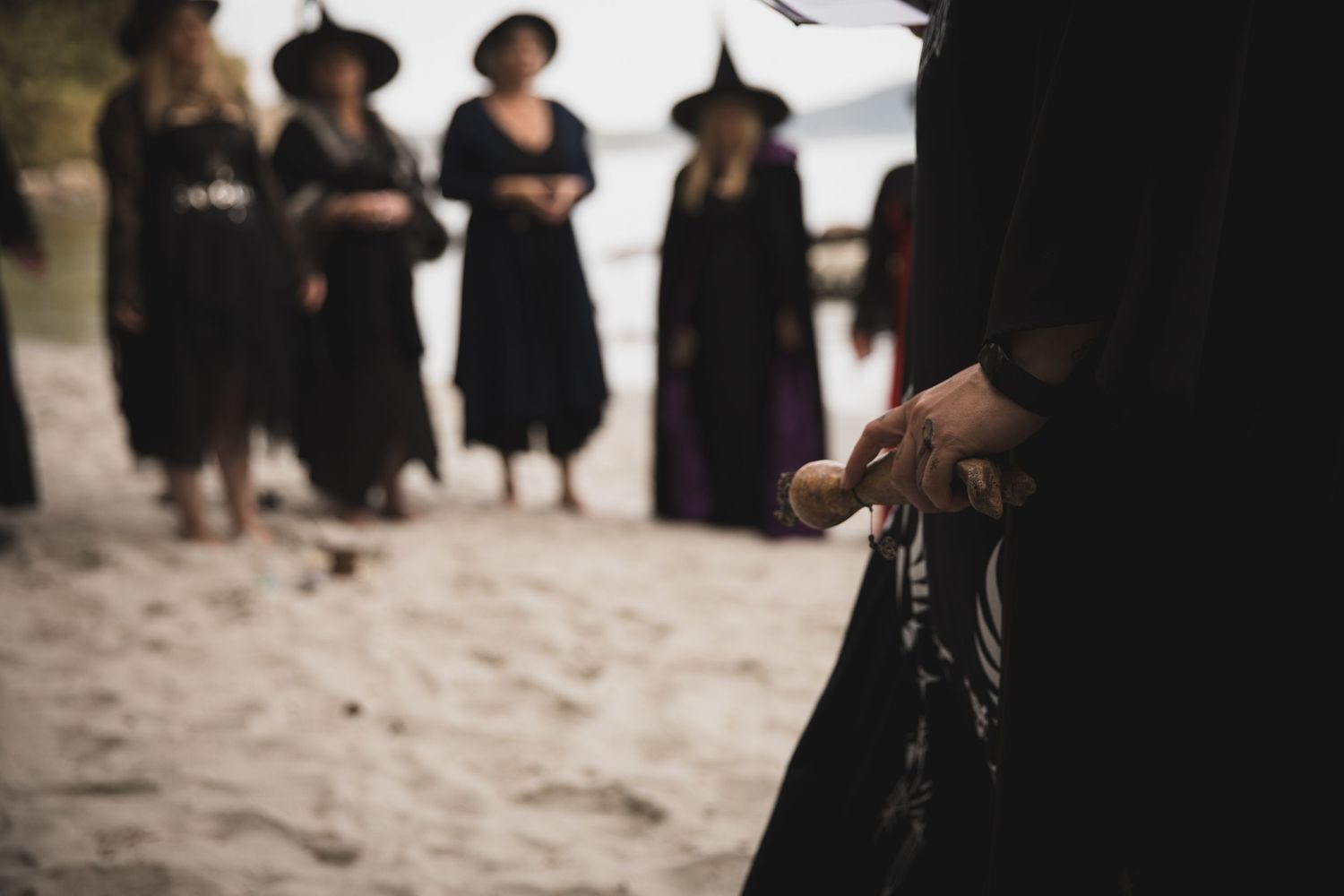 A group of people wearing black dresses and hats stand on a sandy beach in moody atmospheric lighting.
