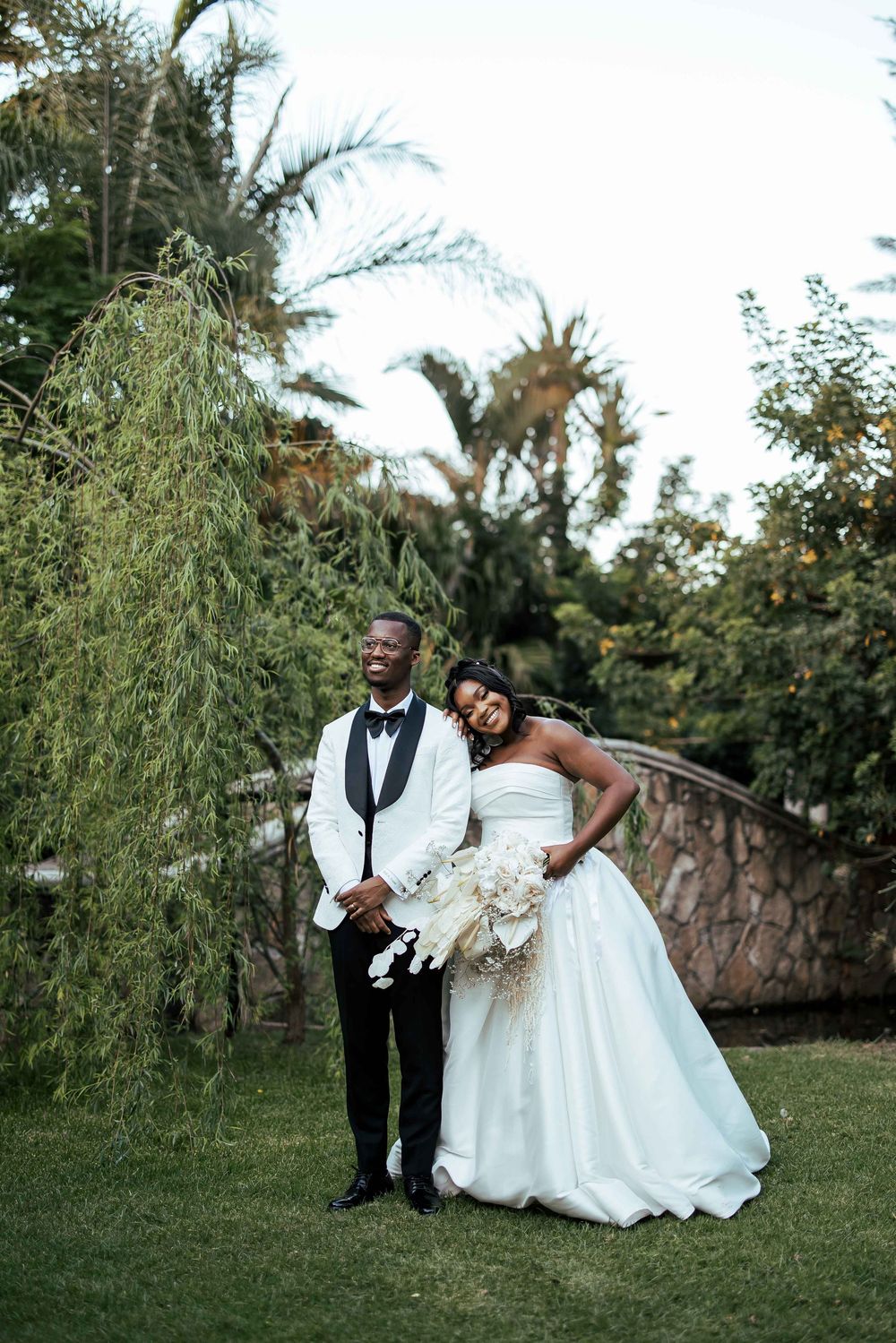 A joyful couple poses together outdoors on their wedding day surrounded by lush greenery and trees.