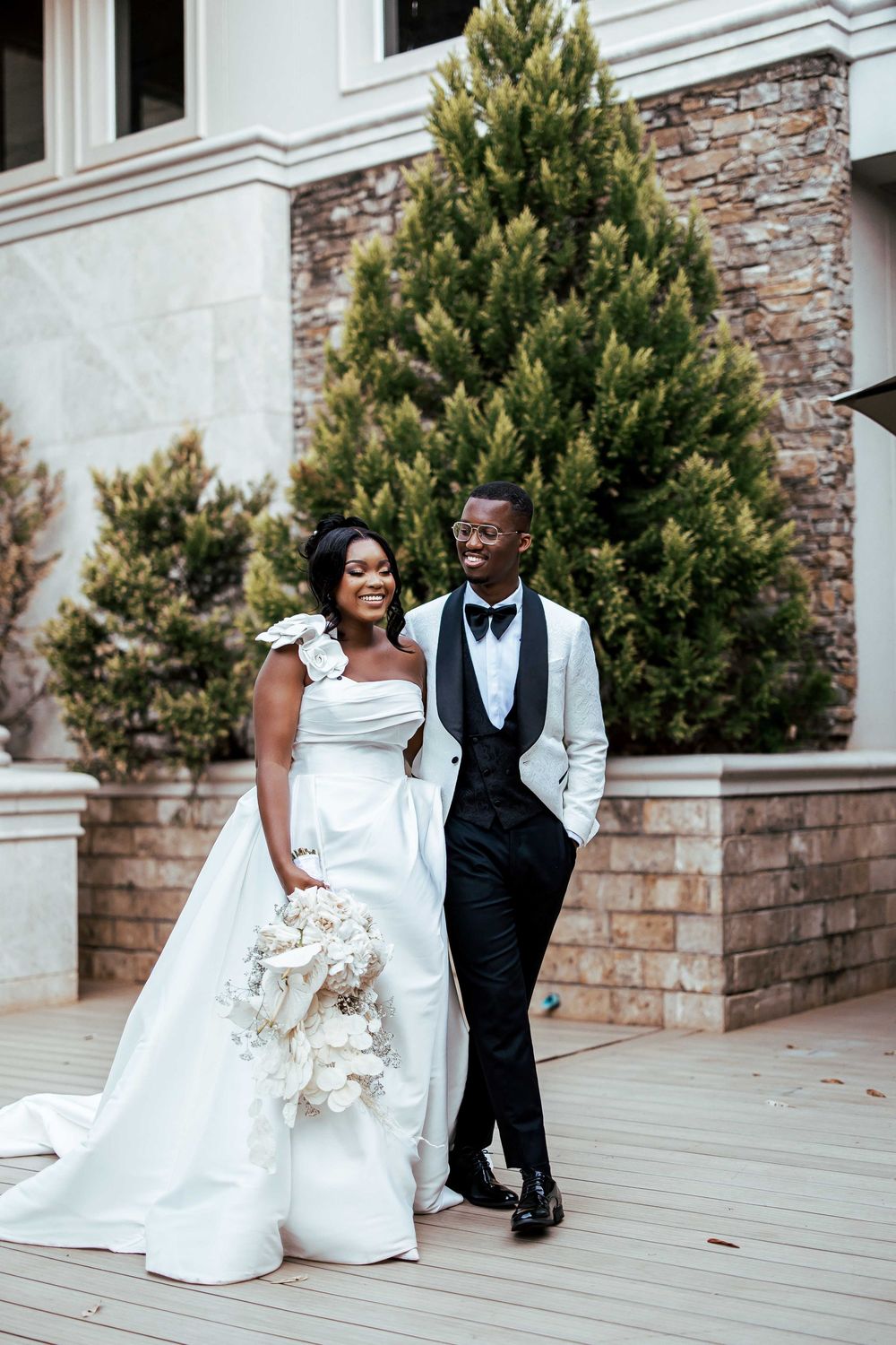 A couple in formal wedding attire shares romantic moments on a stone patio with evergreen trees in the background.