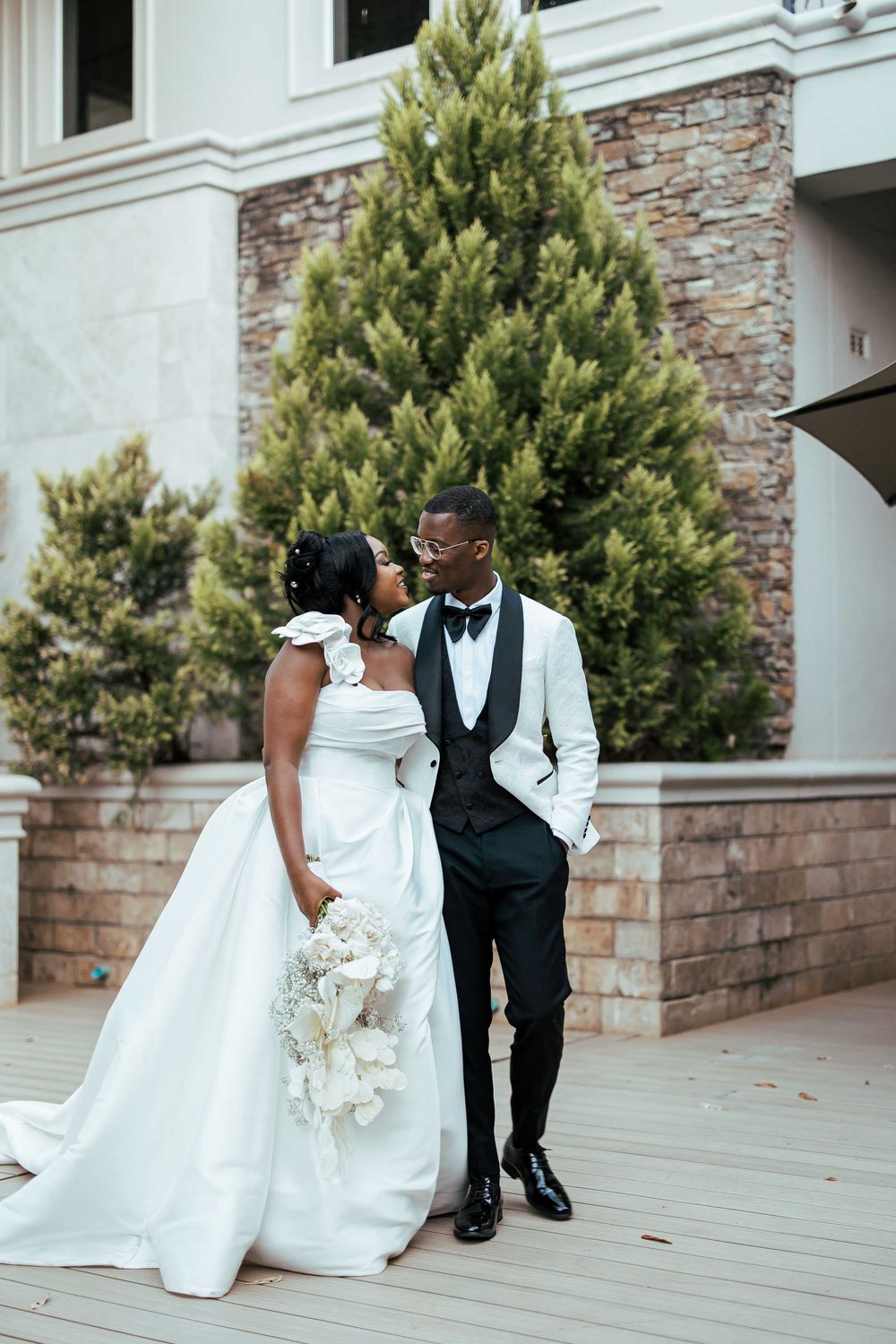 Elegant wedding couple poses together on stone patio with evergreen trees in the background.
