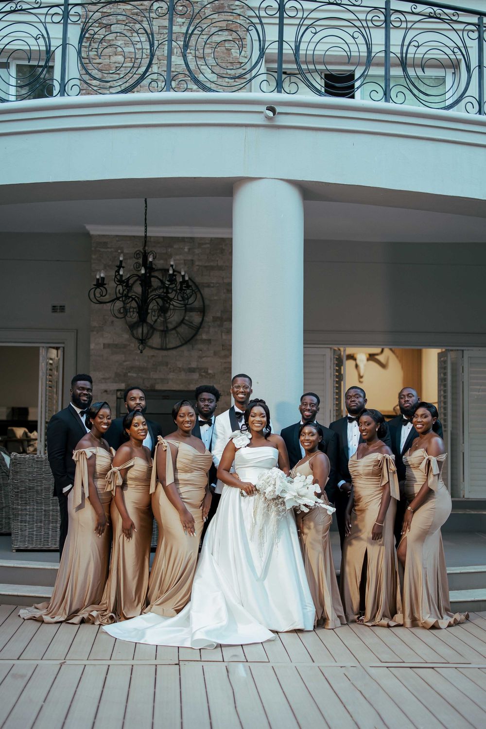 Wedding party in champagne-colored bridesmaid dresses poses with bride in white gown on elegant columned porch.