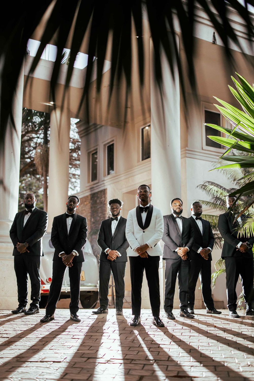 Groomsmen in black tuxedos stand in a line casting dramatic shadows on a tiled floor with palm fronds overhead.