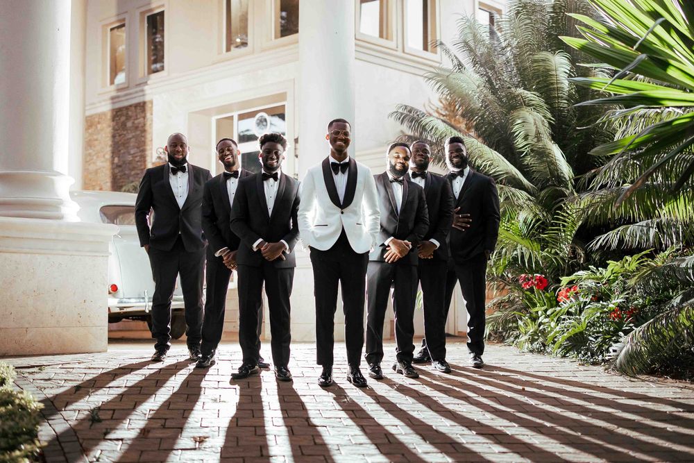 Groomsmen in formal black suits and ties pose together on outdoor steps with palm trees in the background.