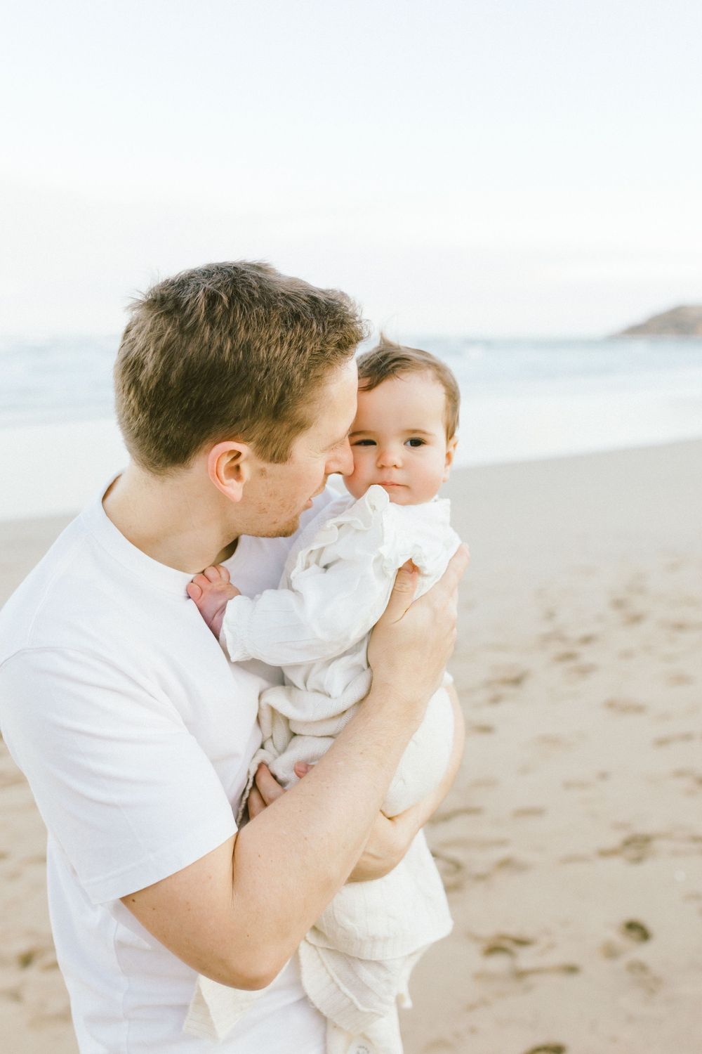 A tender moment between parent and baby in white clothing on a sunny beach.