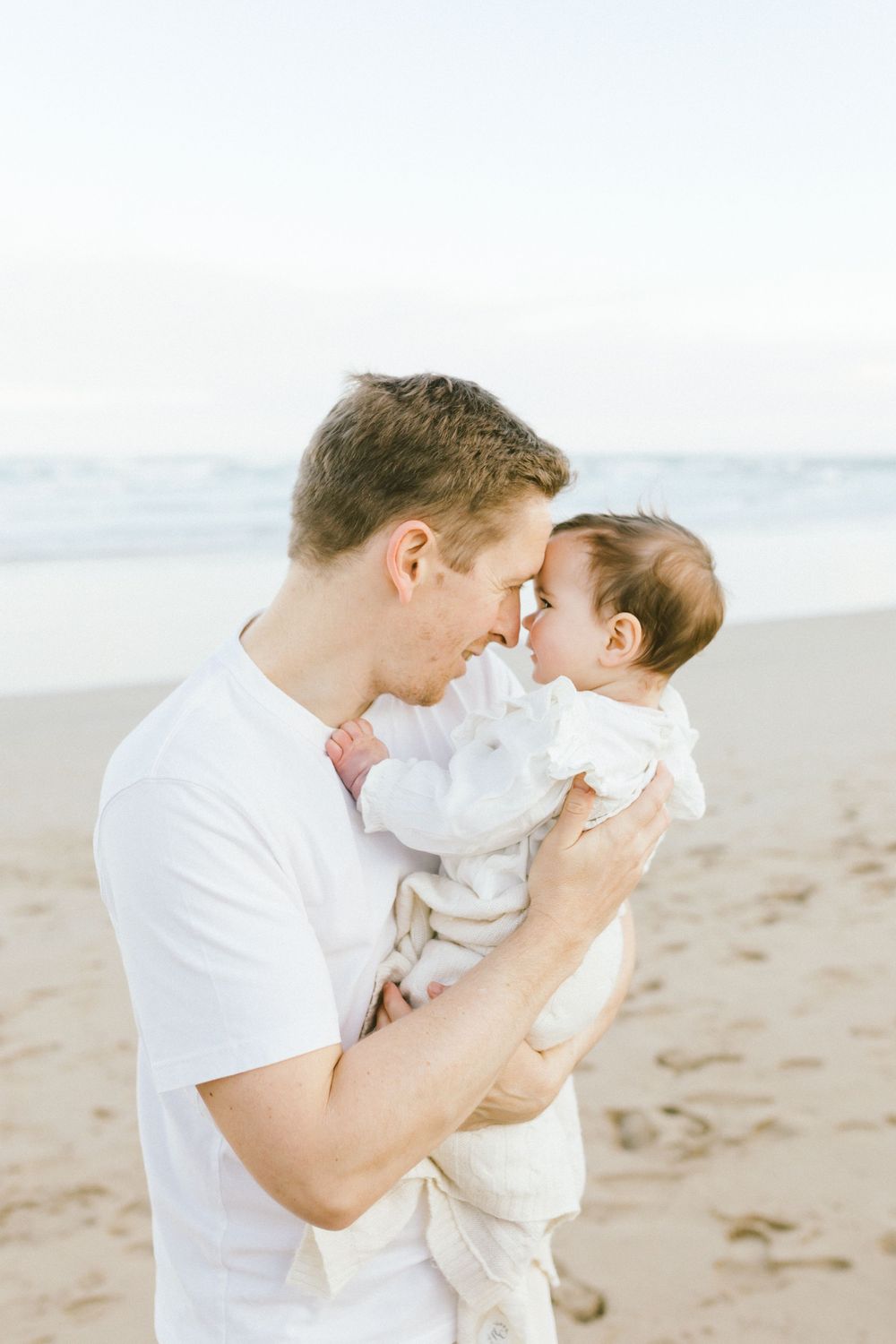 Parent and baby in white clothing share tender moments while playing on a sandy beach at sunset.