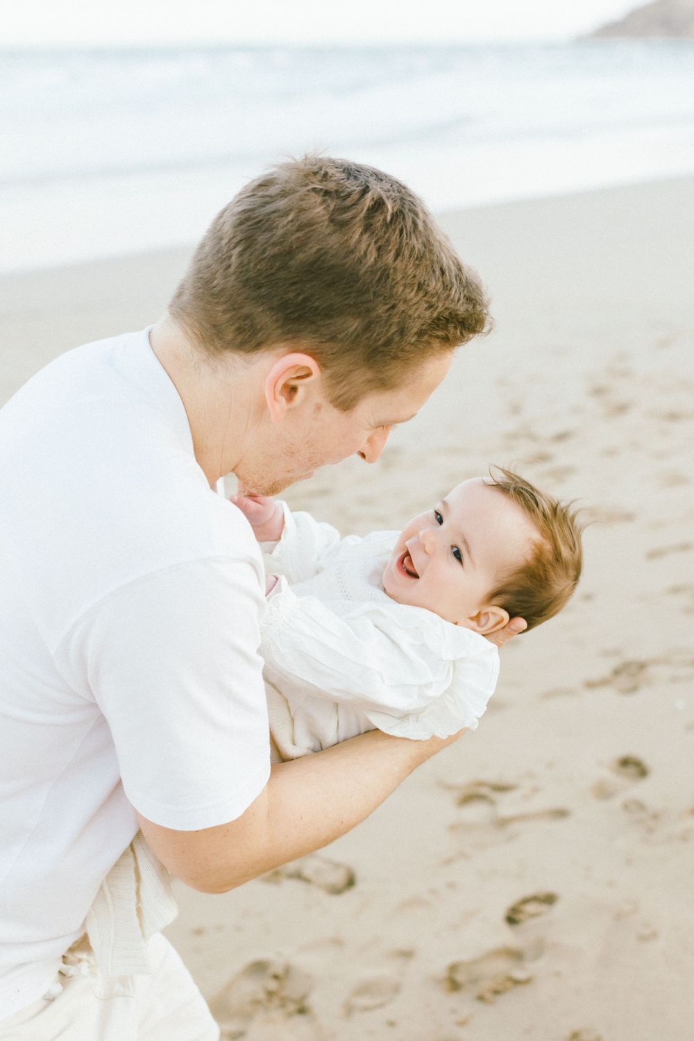 A tender moment captured between parent and child playing together on a sandy beach in white clothing.
