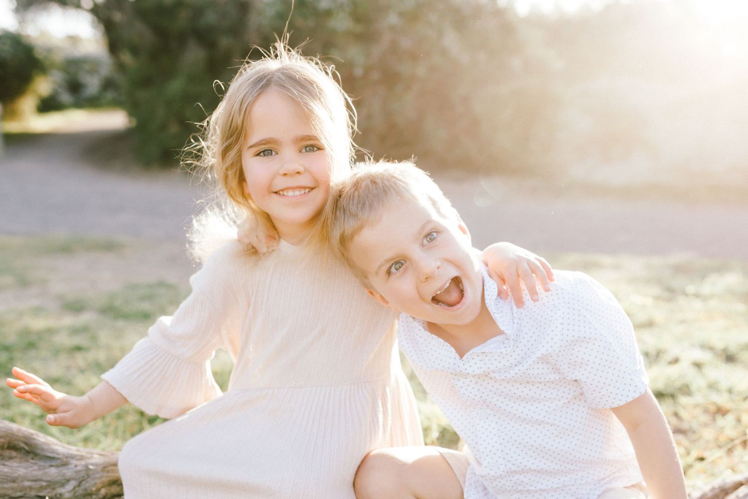 Two people in white outfits embrace while sitting in a sunlit outdoor setting during golden hour.