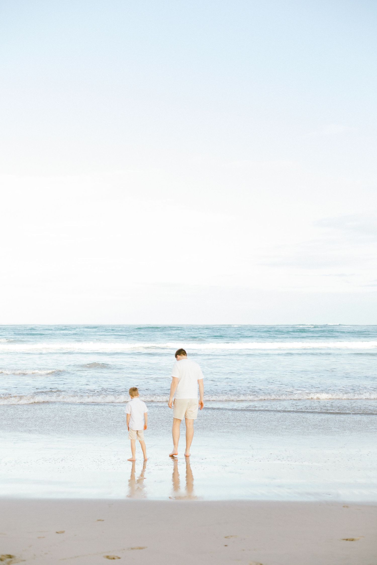 Two figures in white clothing stand at the edge of the ocean waves on a serene beach under a pale blue sky.
