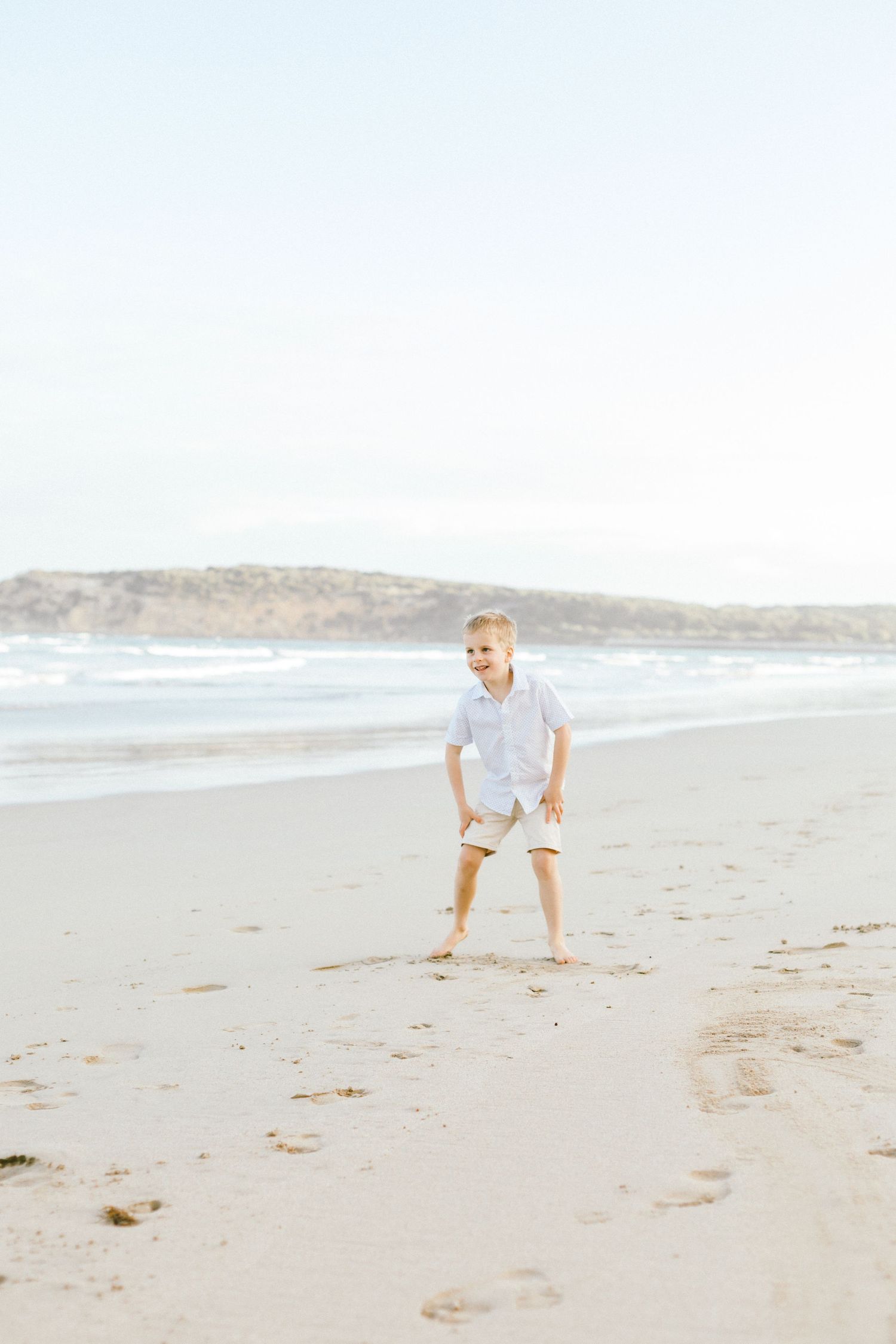A young child in white clothing plays and crouches on a sandy beach with ocean waves visible in the background.