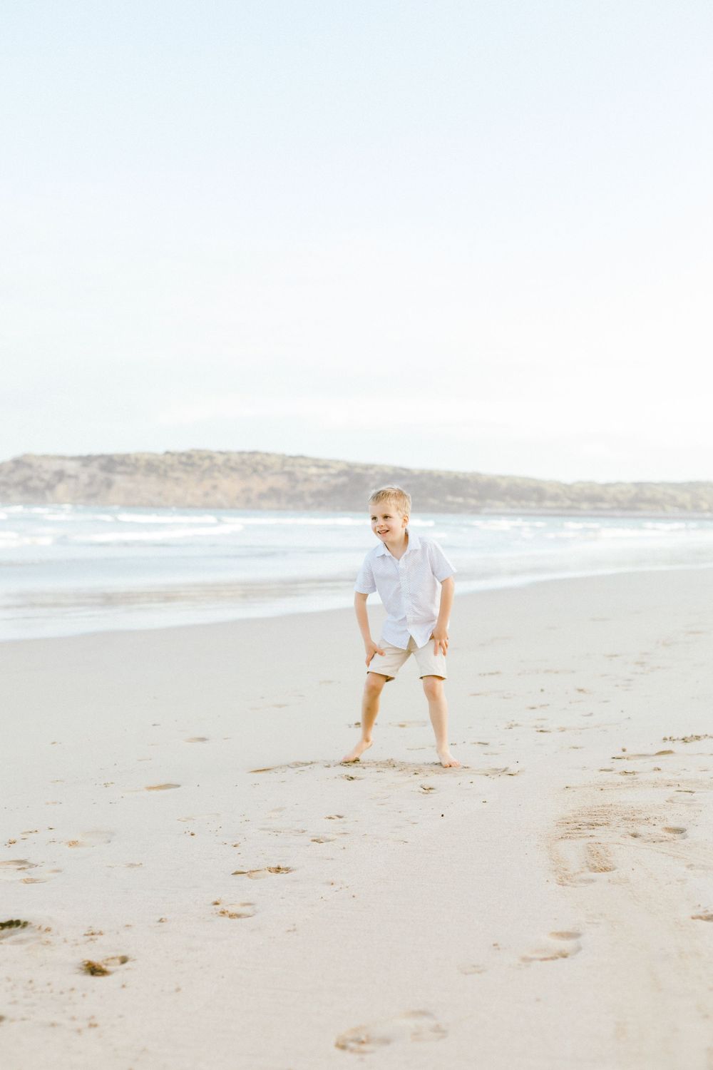 A young child in white clothing plays and crouches on a sandy beach with ocean waves visible in the background.