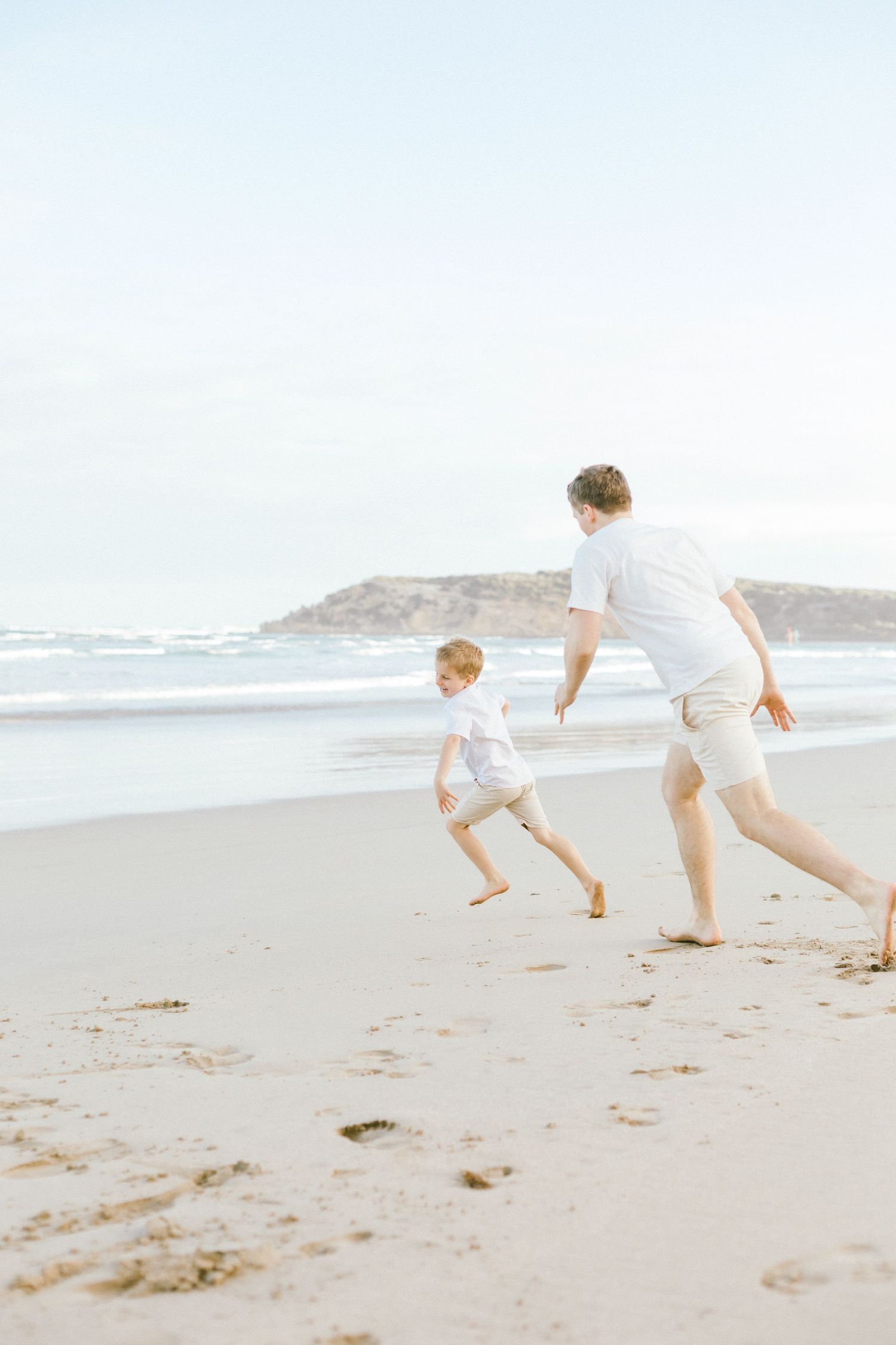 Two people run playfully along a misty beach shoreline on an overcast day.