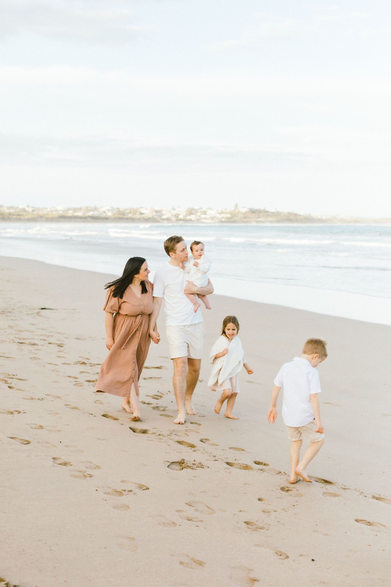 A family walks together on a sandy beach during a sunny day with ocean waves in the background.
