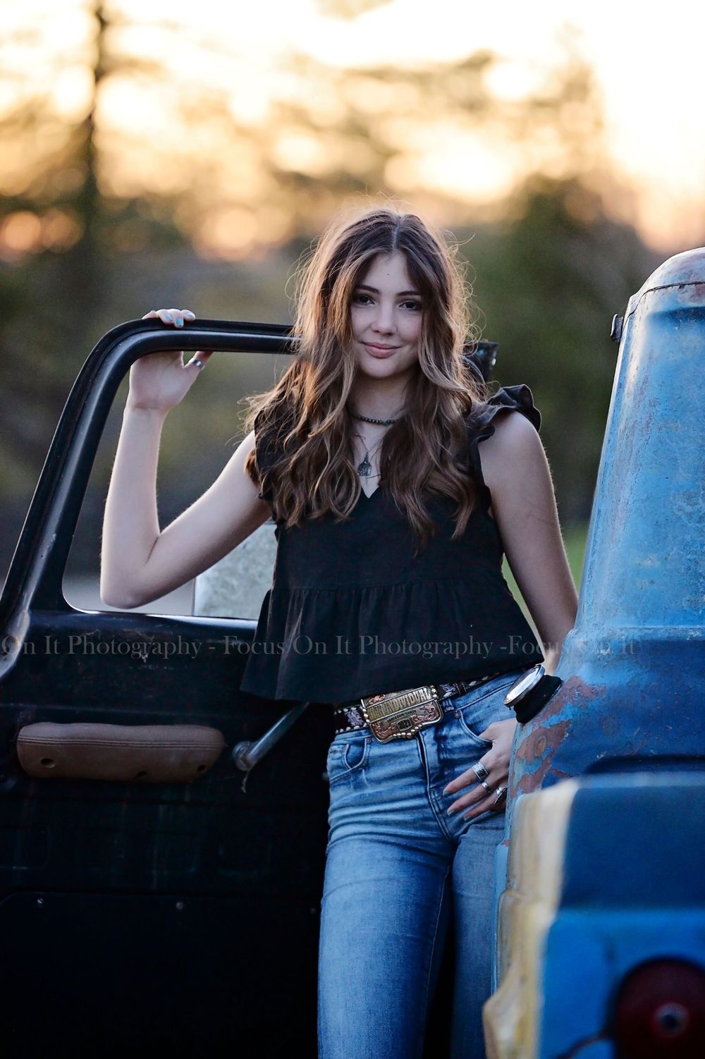 Young woman poses with vintage blue pickup truck during outdoor photo shoot at golden hour.