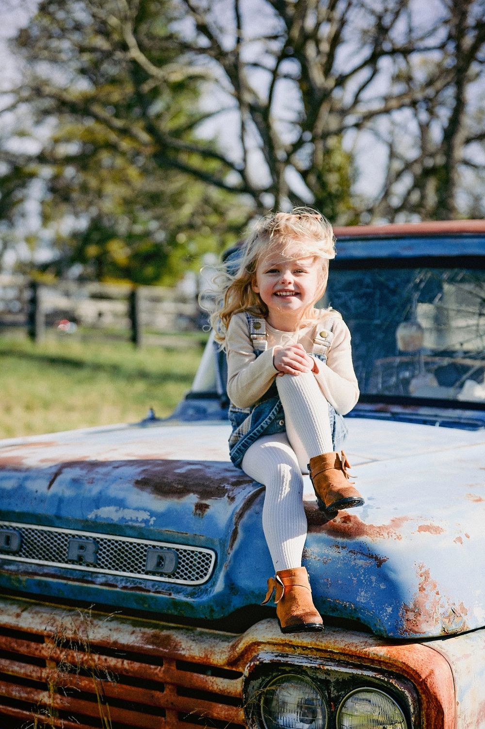Child sitting on hood of vintage blue car in rustic outdoor setting with brown boots and teddy bear.