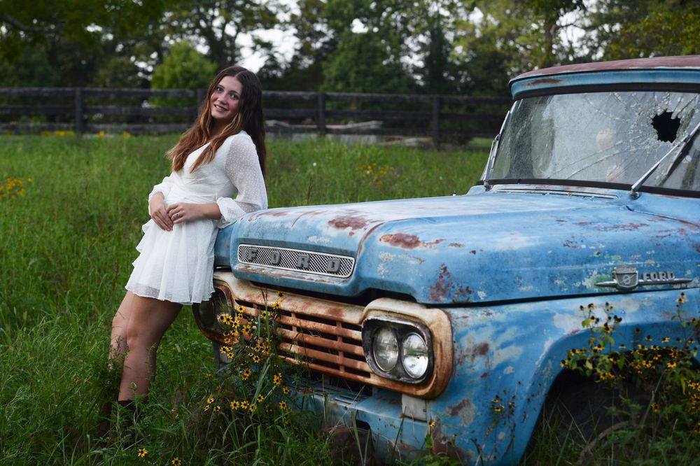 Young woman in white dress poses next to vintage blue Ford pickup truck in grassy meadow during sunset.