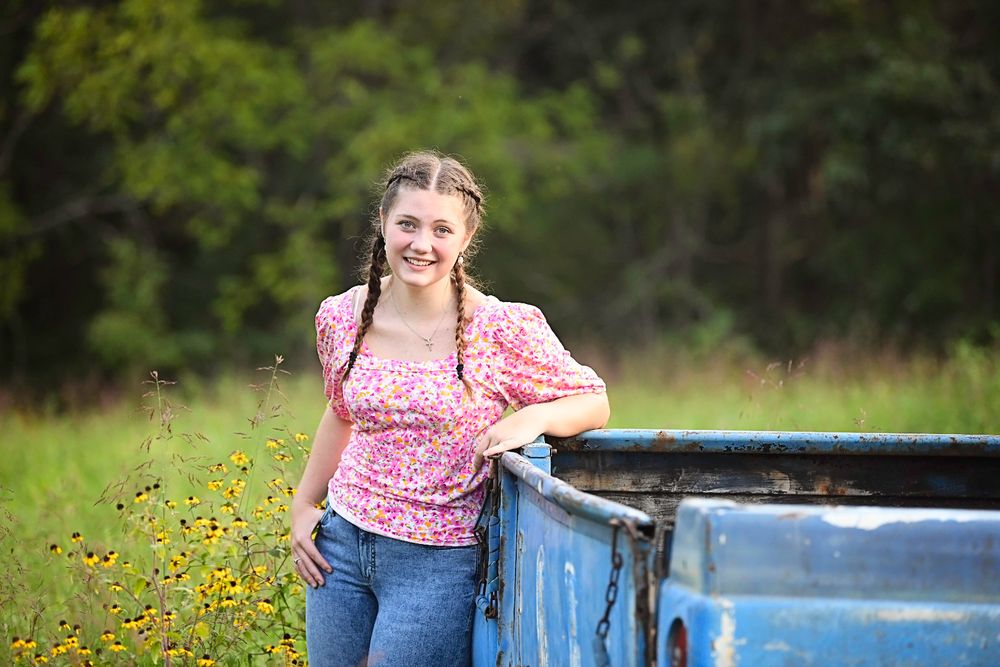 Young person in pink floral top and jeans poses casually against a blue truck with wildflowers in background.