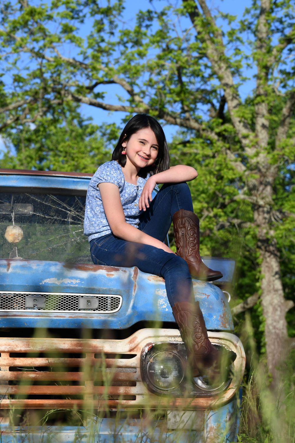 Vintage blue truck hood serves as rustic outdoor portrait setting against green trees on sunny day.