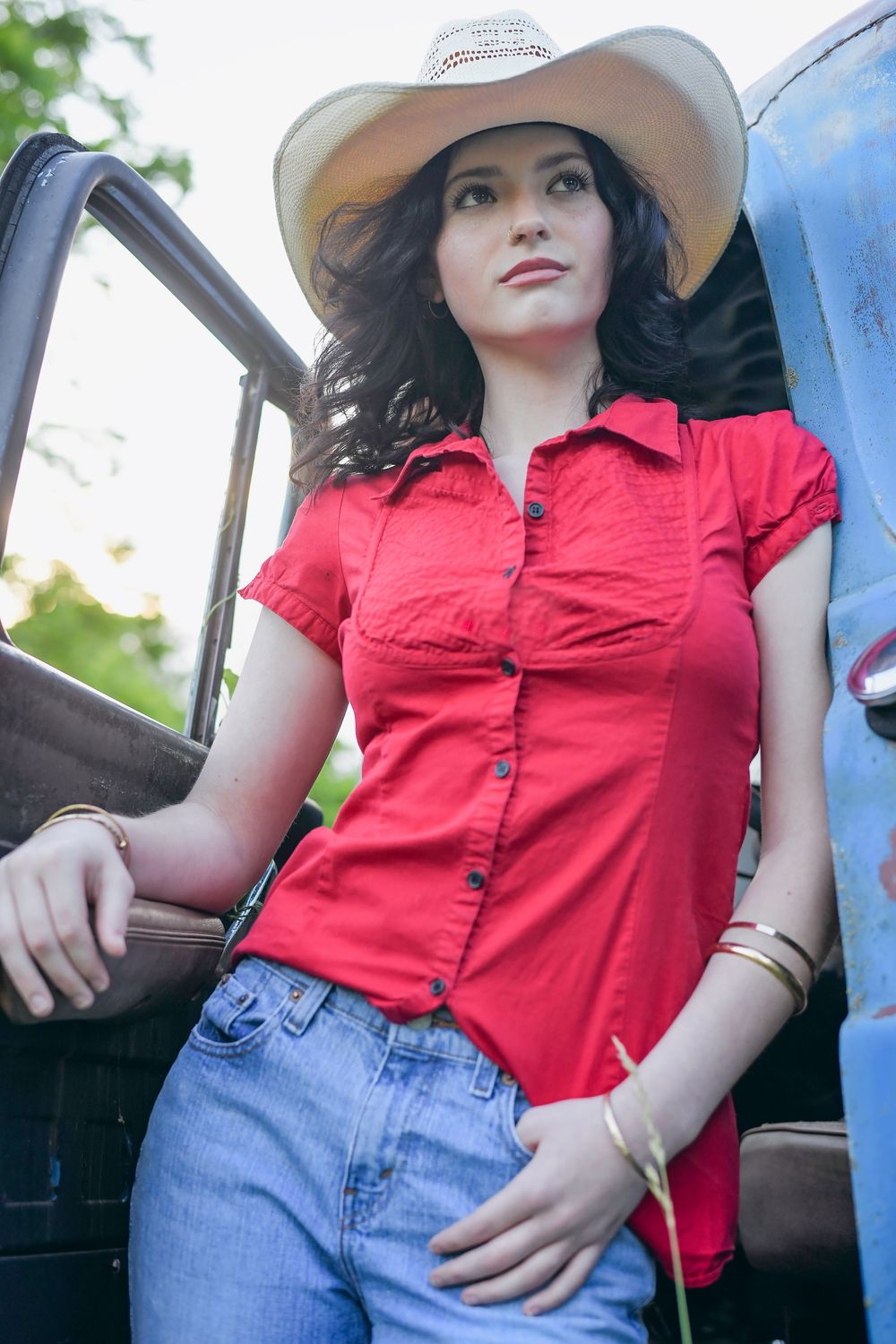 A cowgirl in a red button-up shirt and jeans leans against a blue pickup truck on a sunny day.