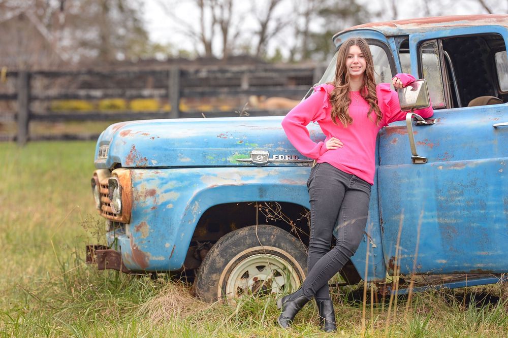 A rustic light blue vintage pickup truck sits in a grassy field as someone in a bright pink sweater leans against it.