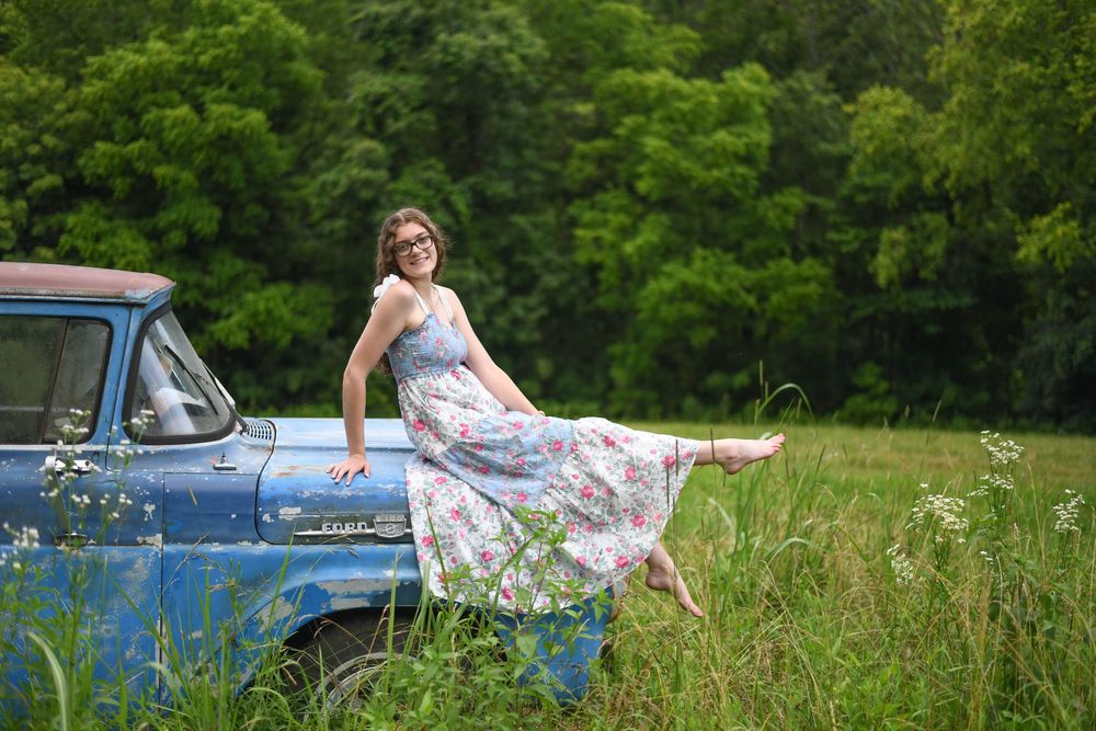 Person in blue dress resting gracefully against vintage car in scenic meadow setting with bright summer lighting.
