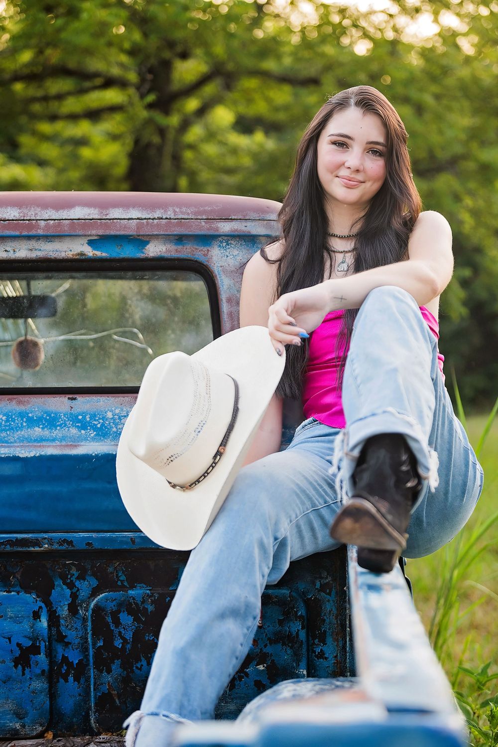 A person in a pink top and jeans sits casually in the bed of a vintage blue pickup truck with a cowboy hat nearby.