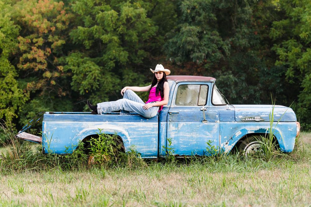 A vintage light blue Ford pickup truck sits in a grassy field with autumn trees in the background.