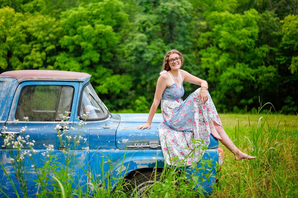 A vintage blue classic car sits in a green field with wildflowers while someone in a floral dress poses on its hood.