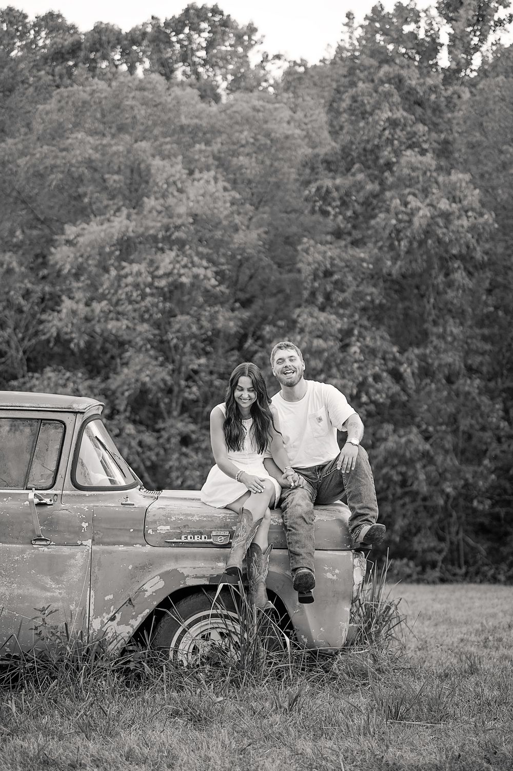 A couple poses together on a vintage car in a rustic outdoor setting with trees in the background.