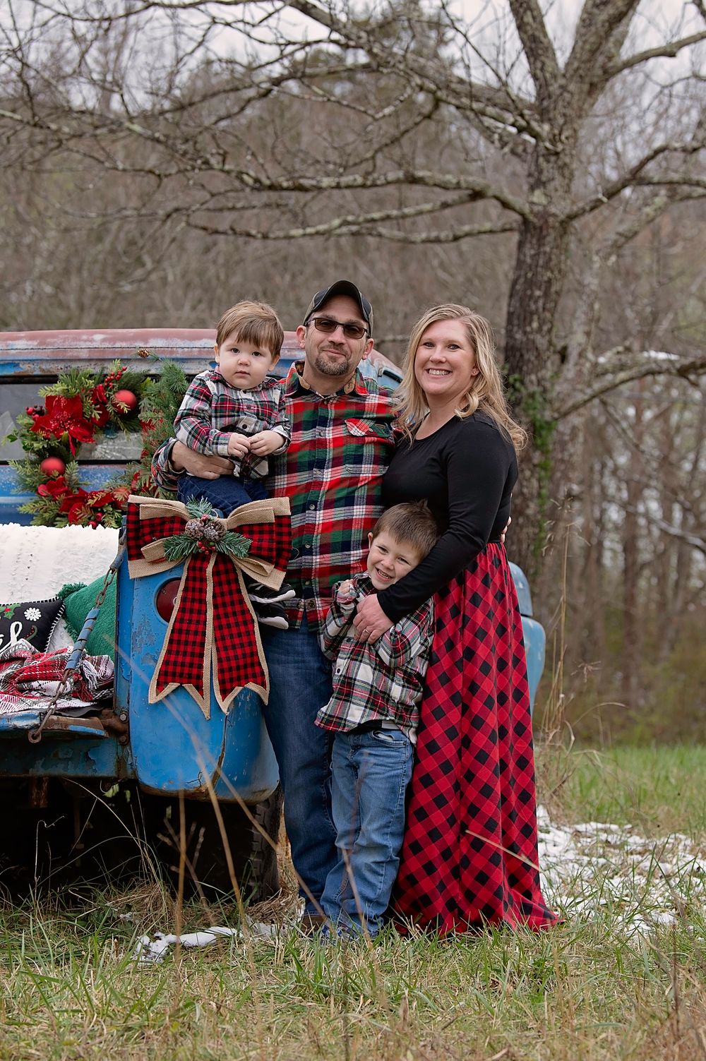 Family poses in plaid outfits for holiday photo by vintage blue truck with Christmas wreath and red bow in rural setting.