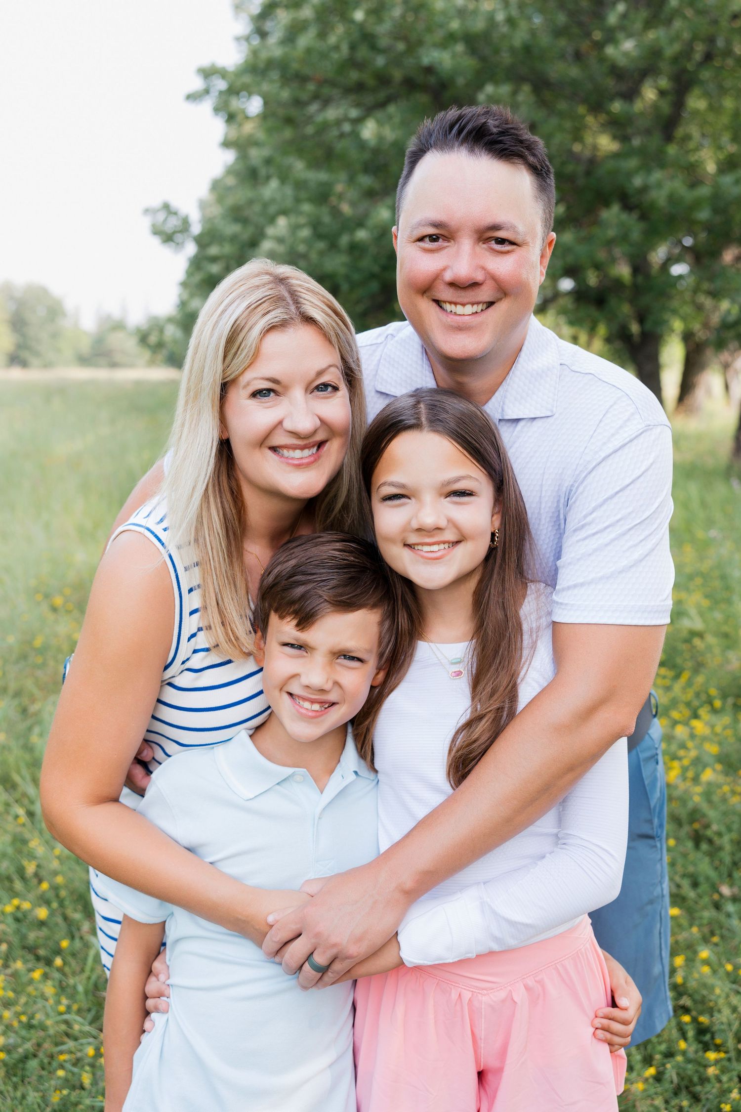 A family smiles together outdoors, dressed in light summer clothing against a natural green background.