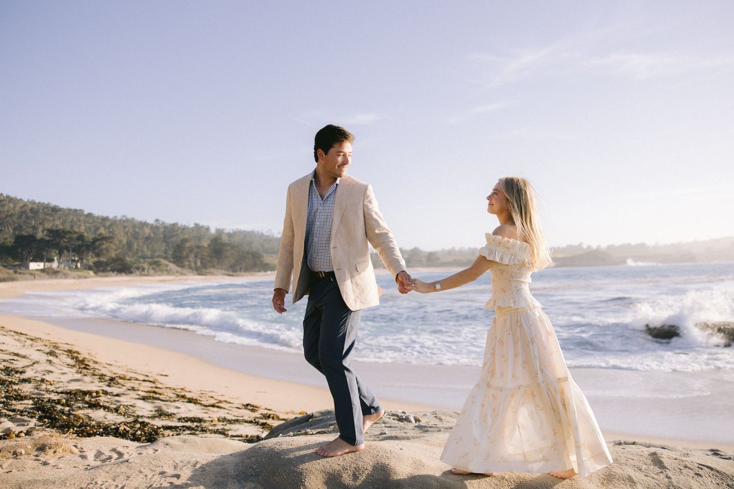 A couple holds hands while walking along a scenic beach shoreline as waves crash during golden hour.