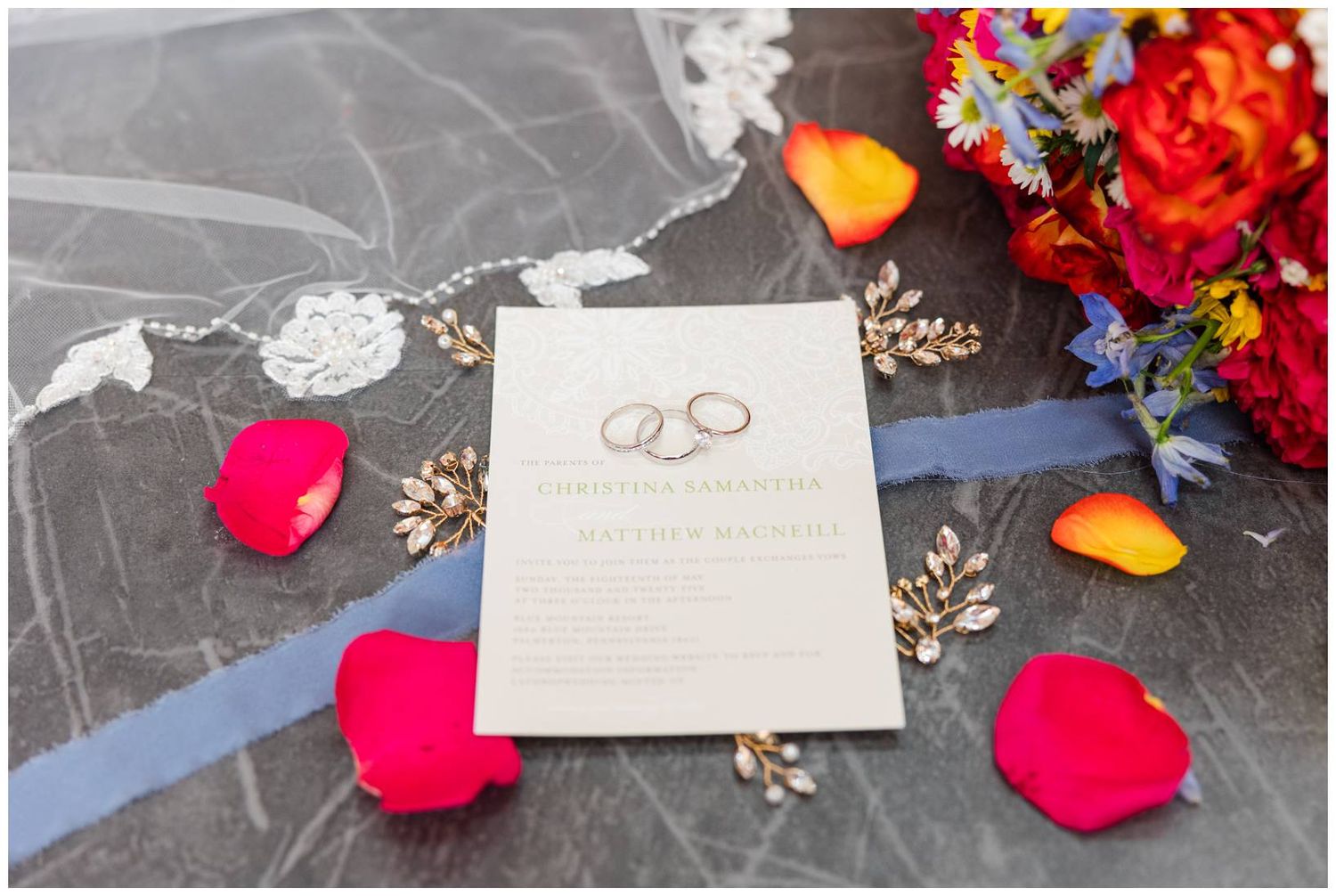 Wedding invitation laid on dark surface with scattered pink rose petals and colorful floral decor in corner.