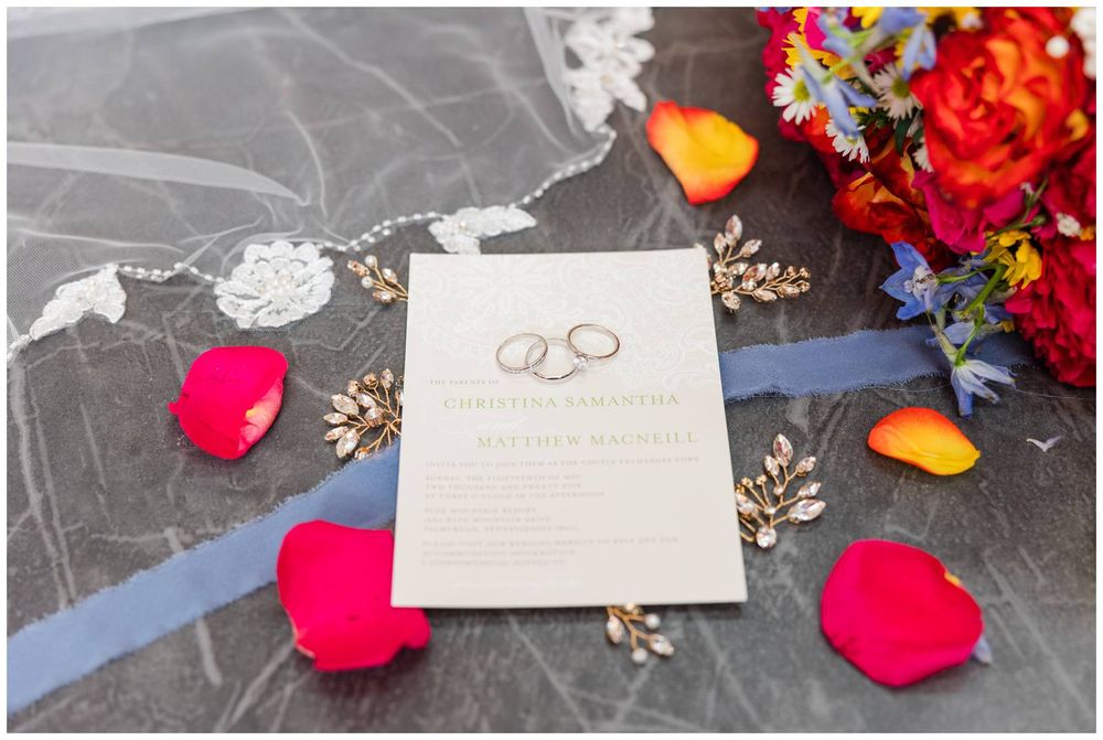 Wedding invitation laid on dark surface with scattered pink rose petals and colorful floral decor in corner.