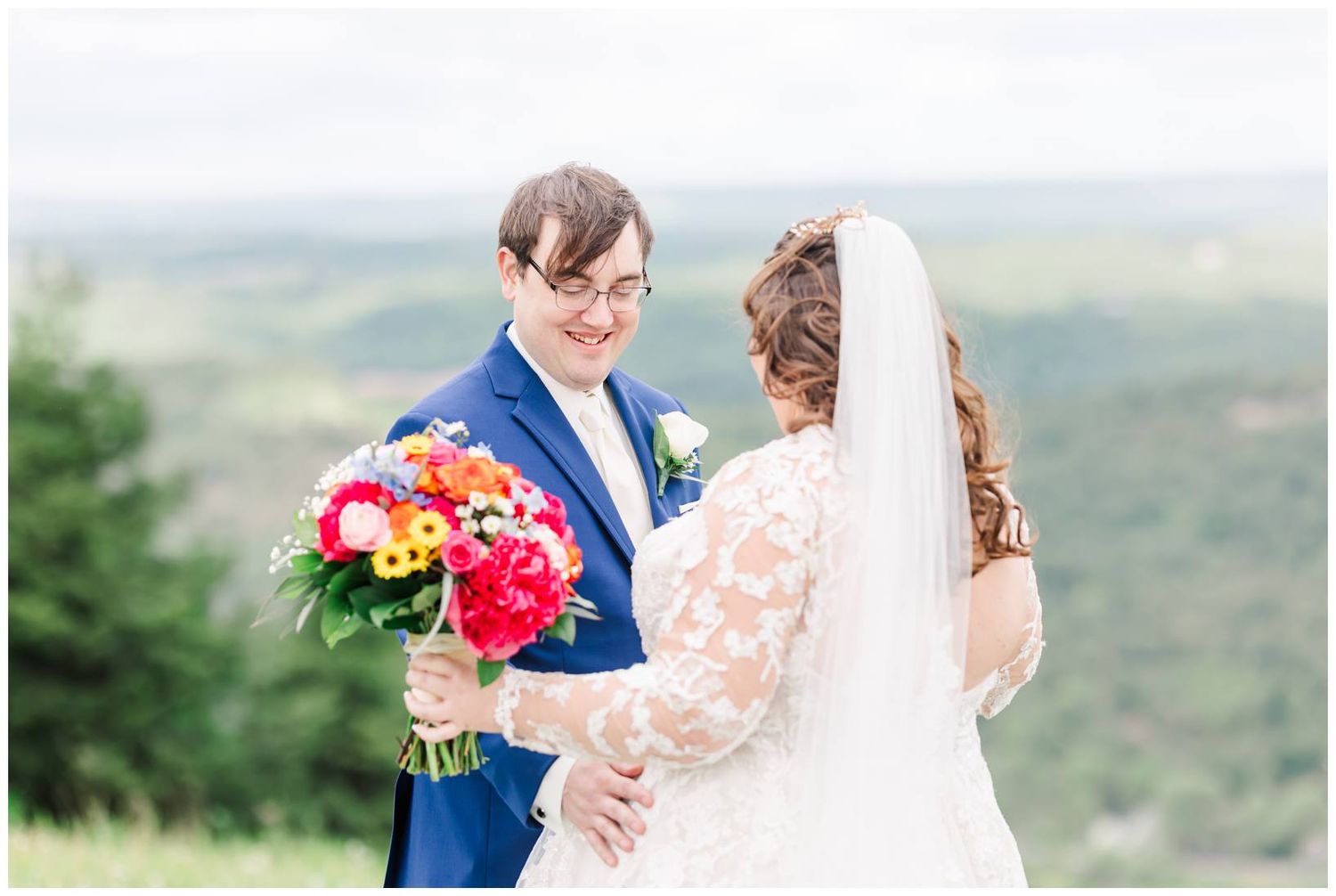 A wedding couple embraces on a scenic hilltop while holding a colorful bouquet of red and orange flowers.