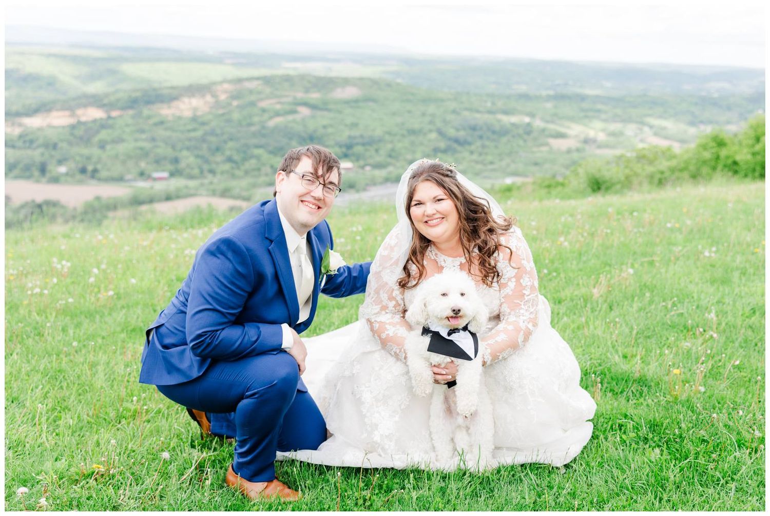 Couple in wedding attire sits on grassy hillside with scenic mountain valley view in background.