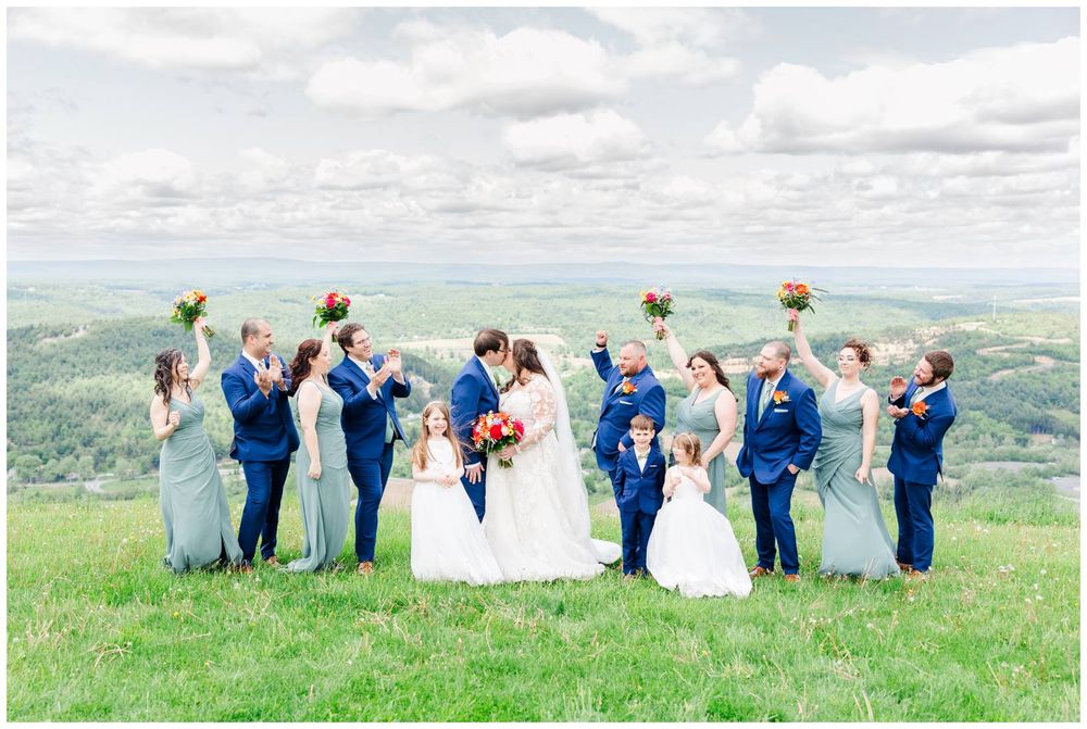 Wedding party celebrates on hilltop meadow with raised arms against scenic mountain backdrop in blue attire.