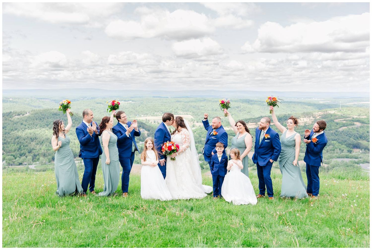 Wedding party celebrates on hilltop meadow with raised arms against scenic mountain backdrop in blue attire.
