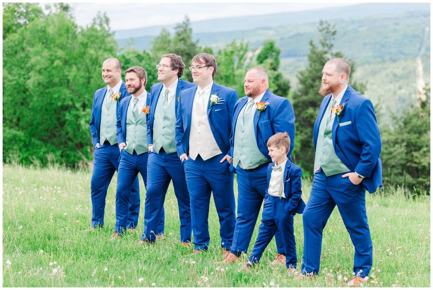 A group wearing matching royal blue suits and sage green ties stands on a grassy hillside overlooking a scenic valley.