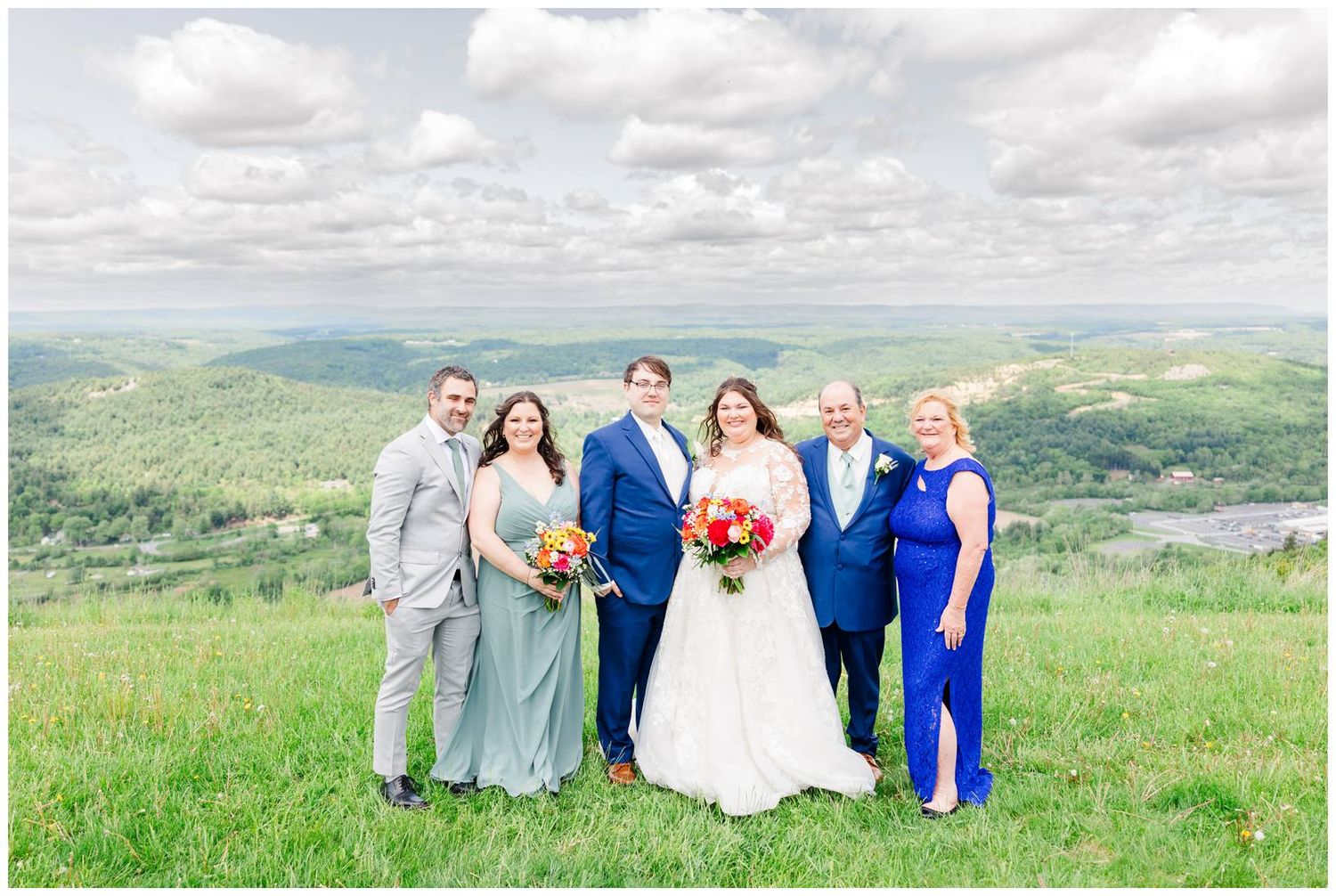 Wedding party poses together on a green hilltop with scenic mountain valley views in the background on sunny day.