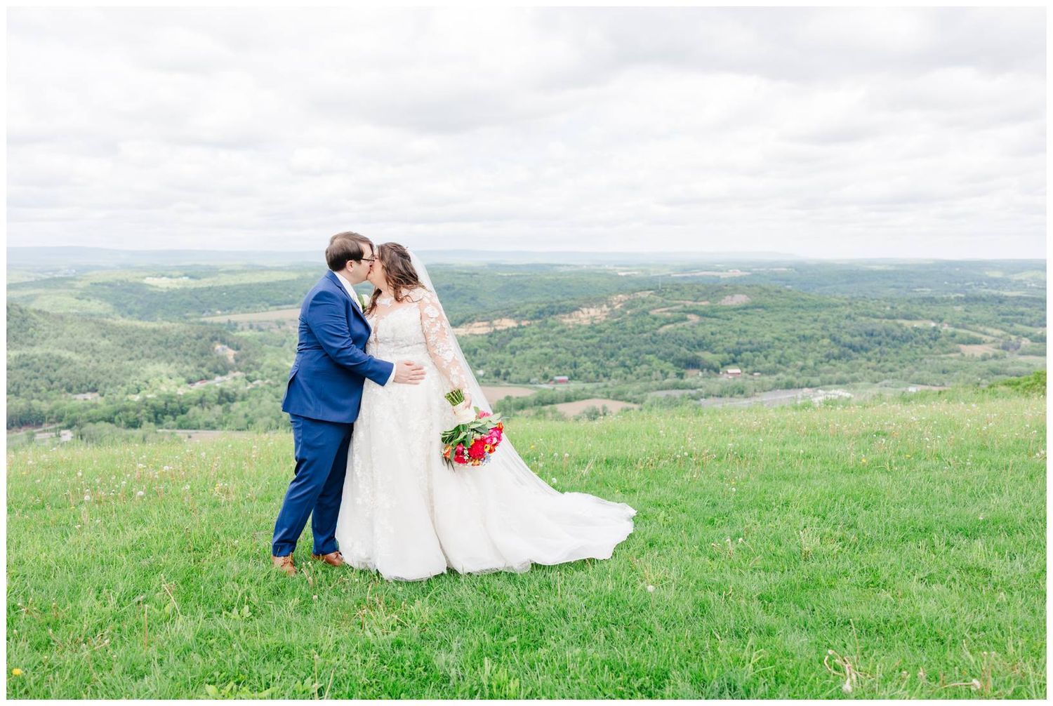A couple in wedding attire share an intimate moment atop a grassy hilltop overlooking a scenic green valley landscape.