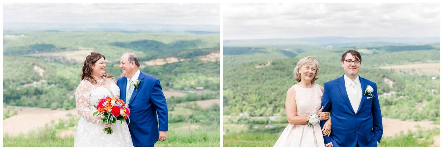Two couples in formal attire stand on a hilltop overlooking a scenic valley landscape on a sunny day.
