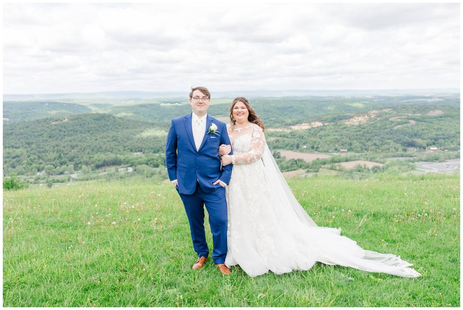 Newlyweds pose together on a grassy hilltop overlooking a scenic valley of rolling green hills and forests.