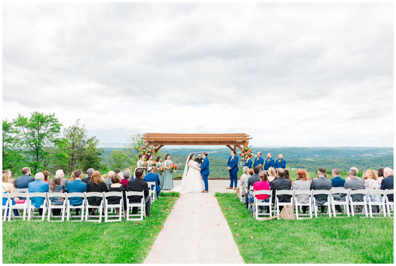 A scenic outdoor wedding ceremony takes place under a wooden arch on a hilltop with guests seated on white chairs.
