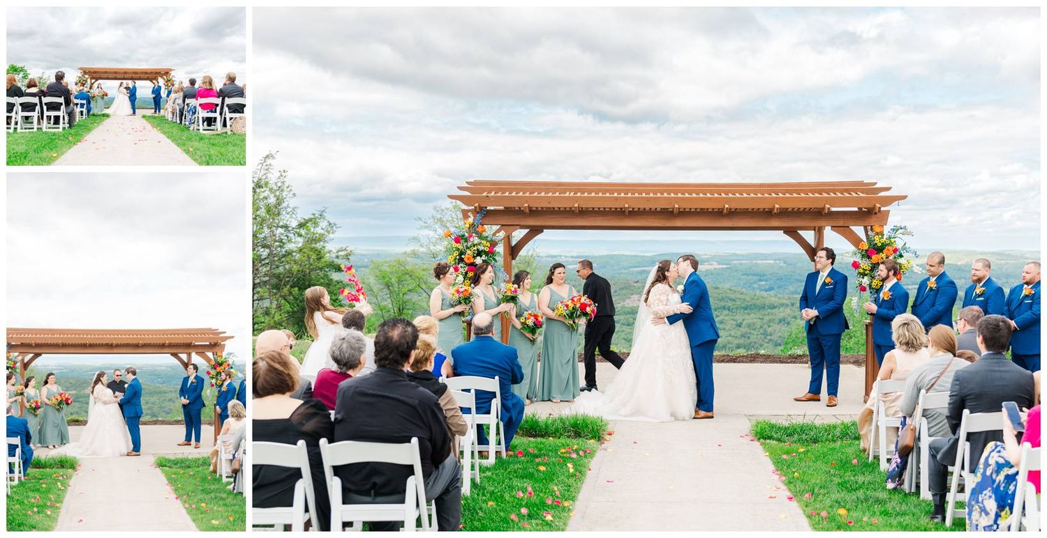 An outdoor wedding ceremony takes place under a wooden pergola on a hilltop with guests seated on white chairs.