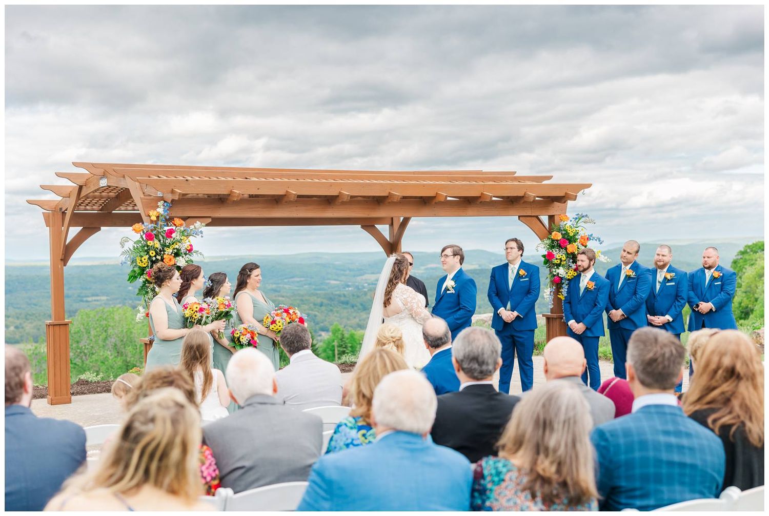 Mountain wedding ceremony takes place under a wooden pergola decorated with flowers with guests seated outdoors.