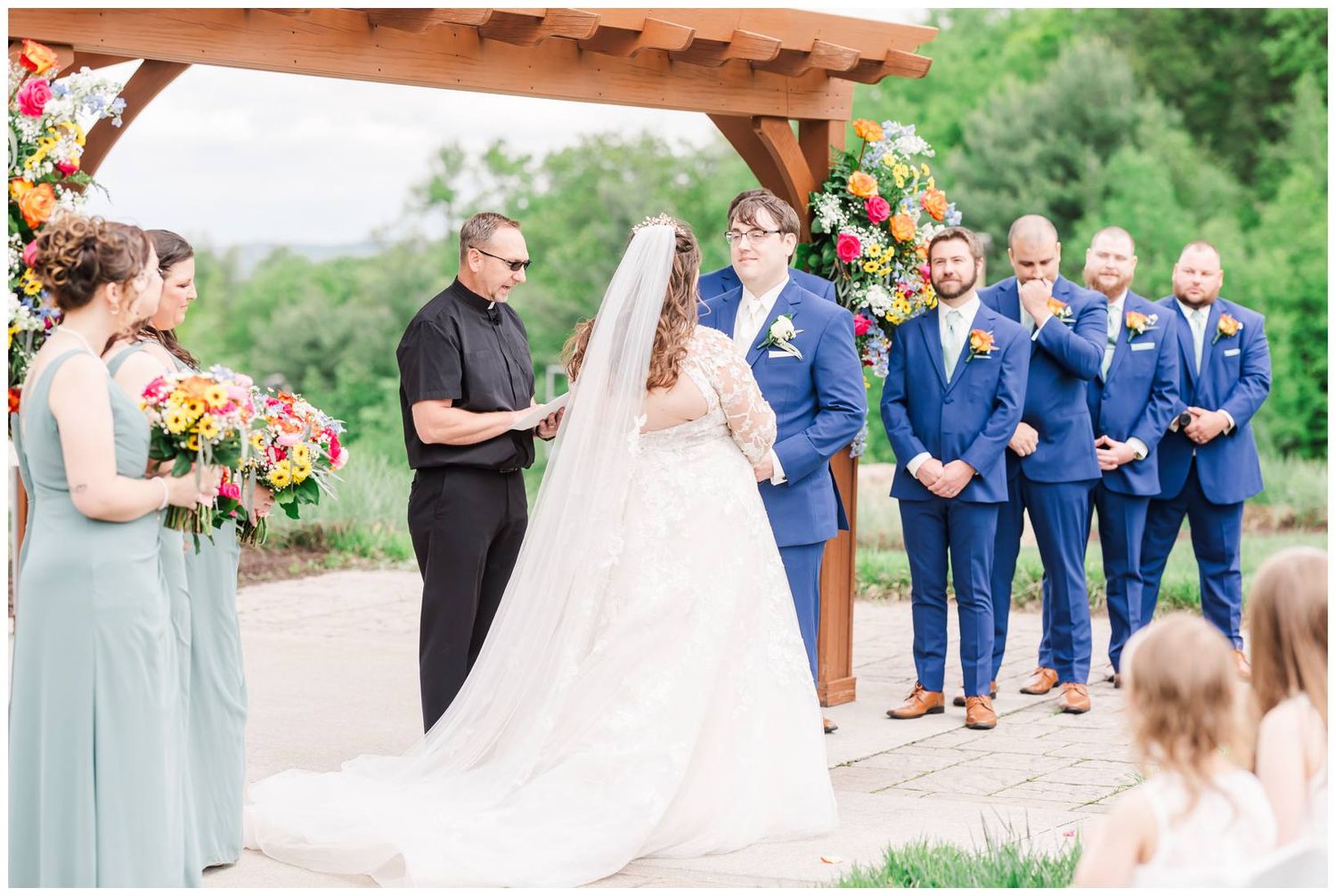 An outdoor wedding ceremony under a wooden pergola with bridesmaids in sage green dresses and groomsmen in navy suits.