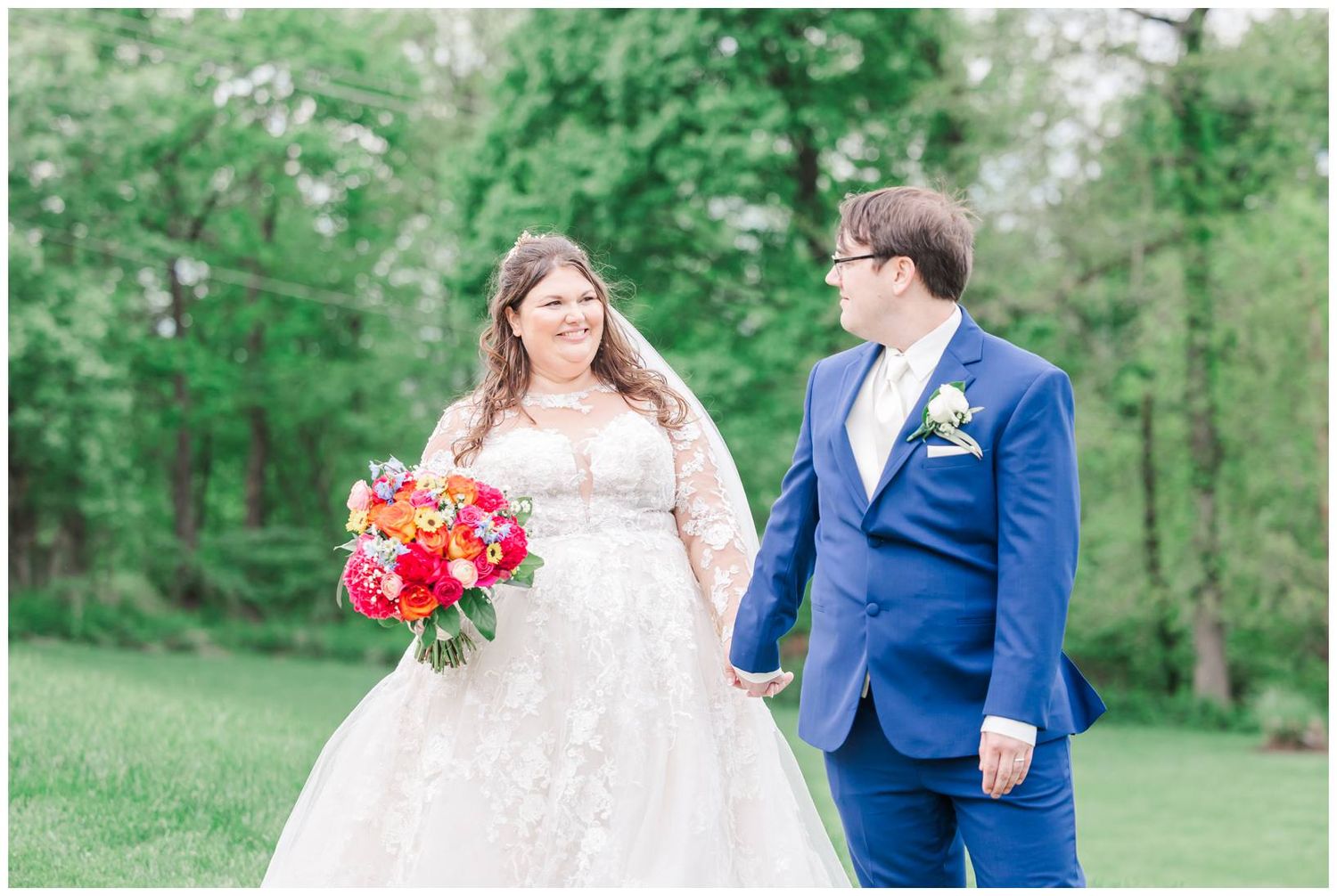 A couple standing together in a green garden setting, the person in white holding a vibrant red bouquet.