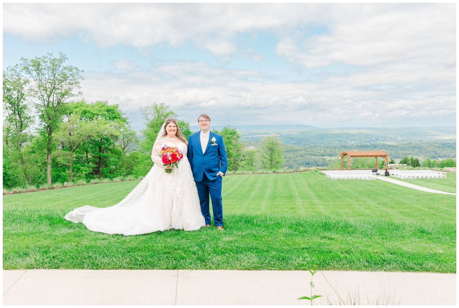A wedding couple stands on a grassy hilltop overlooking a scenic countryside landscape on a cloudy spring day.